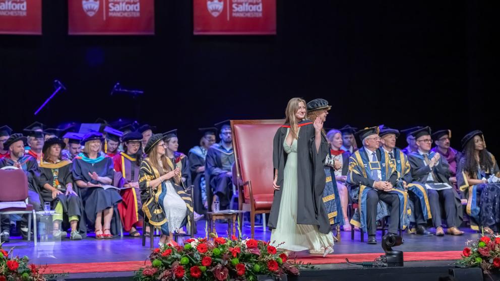 Graduate in a Graduation gown crosses the stage while waving to the audience.