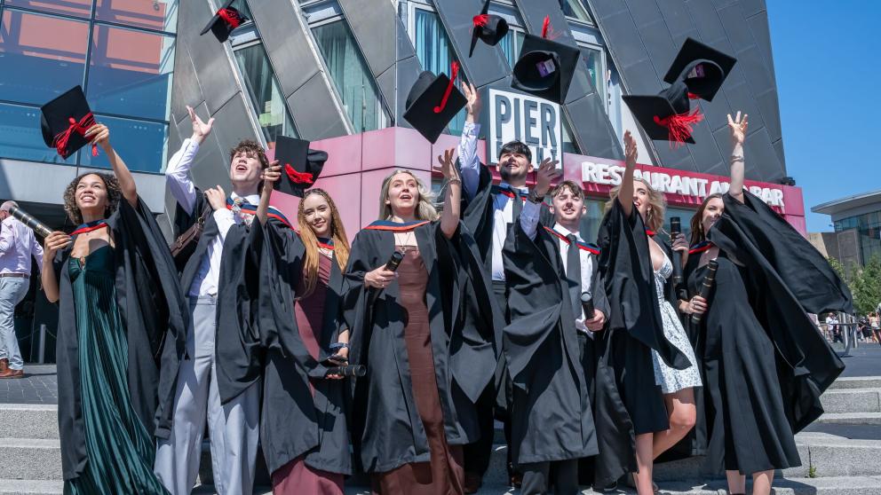 Graduates in Graduation gowns throw their mortar board hats ,with red tassels, in the air. They stand outside the Lowry theatre on a sunny day.