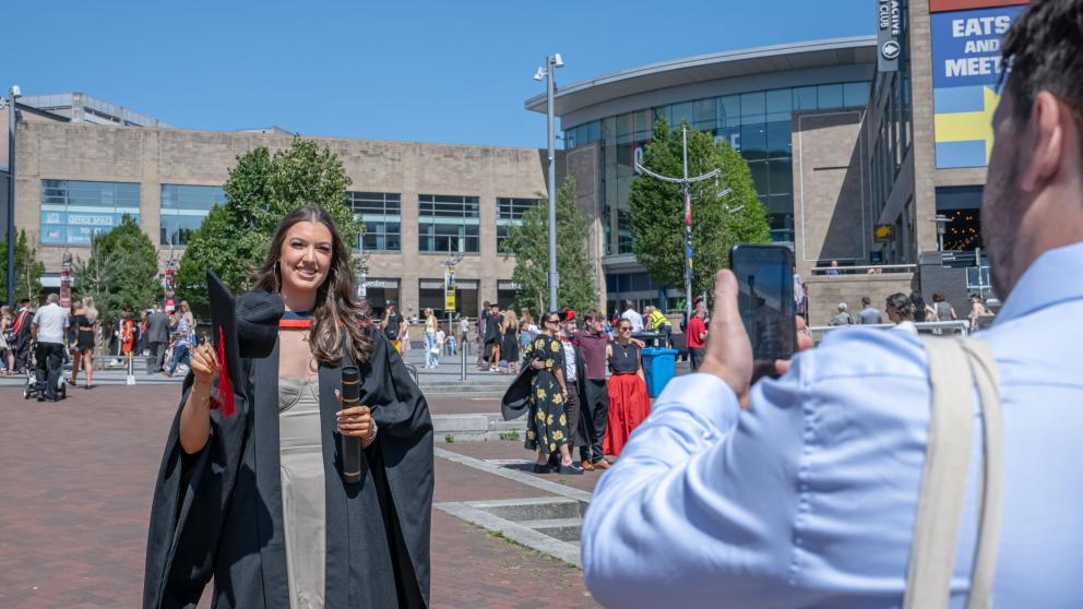 A graduate smiles as their picture is taken using a phone, which can be seen being held in the corner of the picture. The graduate holds a black mortar board hat with a red tassel and wears a black graduation gown on top of a long stone coloured dress.