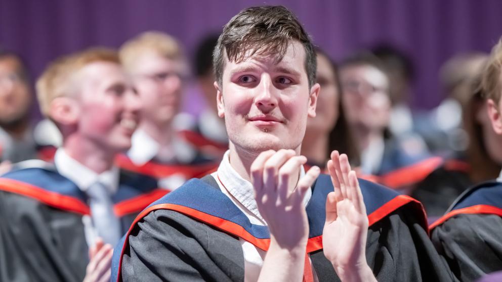 A graduate claps during the ceremony while wearing a black graduation gown with a blue and red hood.