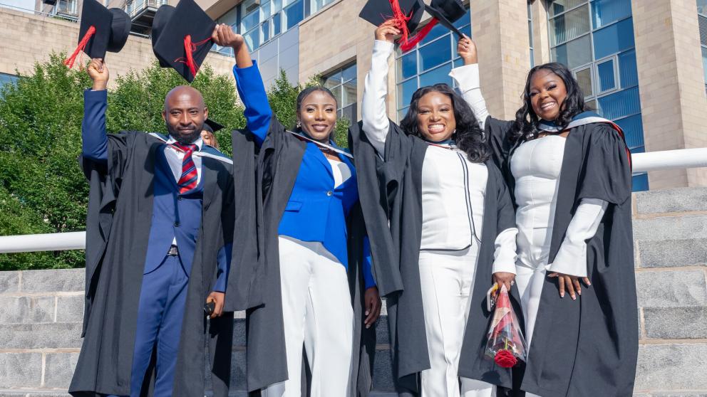 Four graduates stand in their black graduation gowns and lift their caps into the air. The graduates have black mortar board hats with red tassels. They stand outside on a sunny day