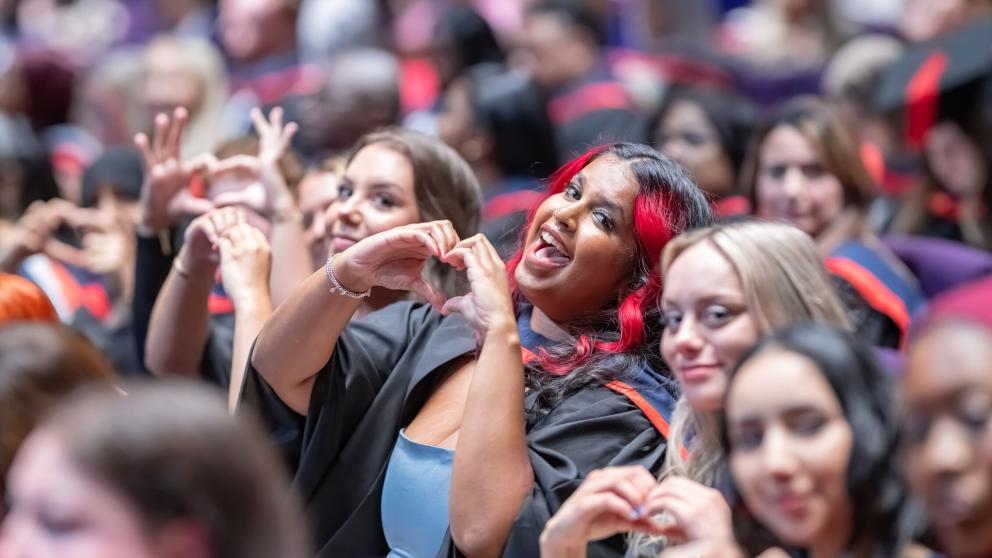 A graduate in the theatre audience makes a heart sign with her hands and smiles at the camera.