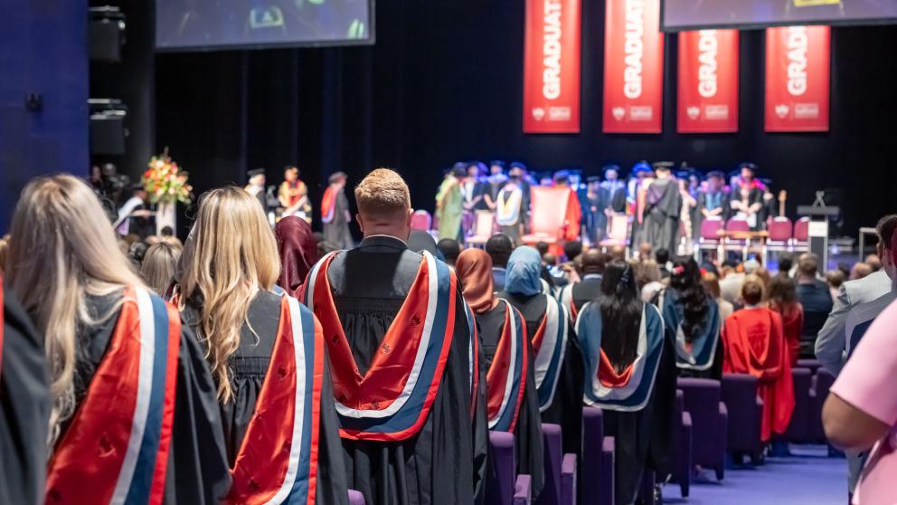 Graduates are photographed from behind stood at the end of their assigned rows in the Lowry Theatre. The graduates wear black graduation gowns with hoods on top. The hoods are dark blue and grey on the outside, and red in the middle with the University of Salford logo repeated over the red in black.