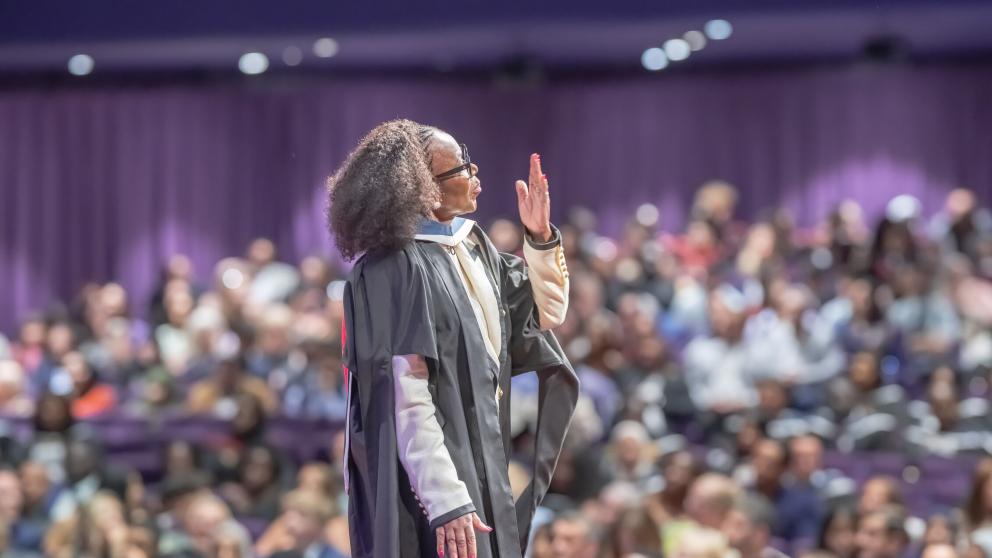 A graduate is photographed from the back. They blow a kiss to the audience, which can be seen in a blur behind them. The graduate wears a black graduation robe. 