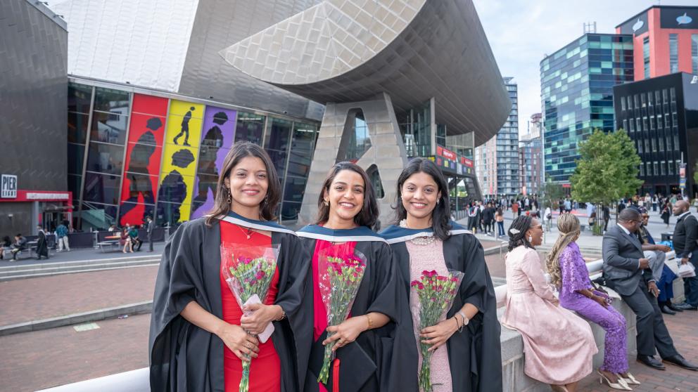 three graduates in Graduation gowns hold bouquets of red flowers and smile at the camera while standing in front of the Lowry theatre.