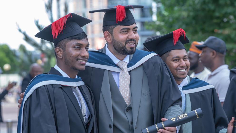 Three graduates in suits and black graduation gowns smile. All wear black mortar board hats with red tassels. The middle graduate holds a black scroll with a gold University of Salford logo on it. 