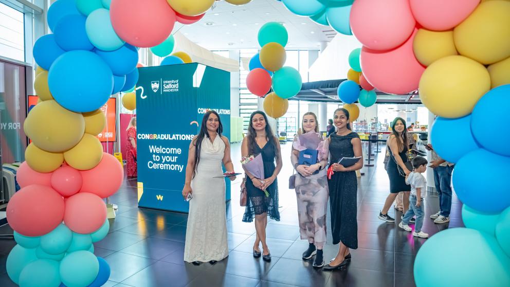 Four graduates in dresses smile under a balloon that is blue, red, teal and yellow. The two center graduates hold flower bouquets and the other two hold Graduation brochures. A cuboid screen in the background reads, 'Congradulations. Welcome to your Ceremony.'