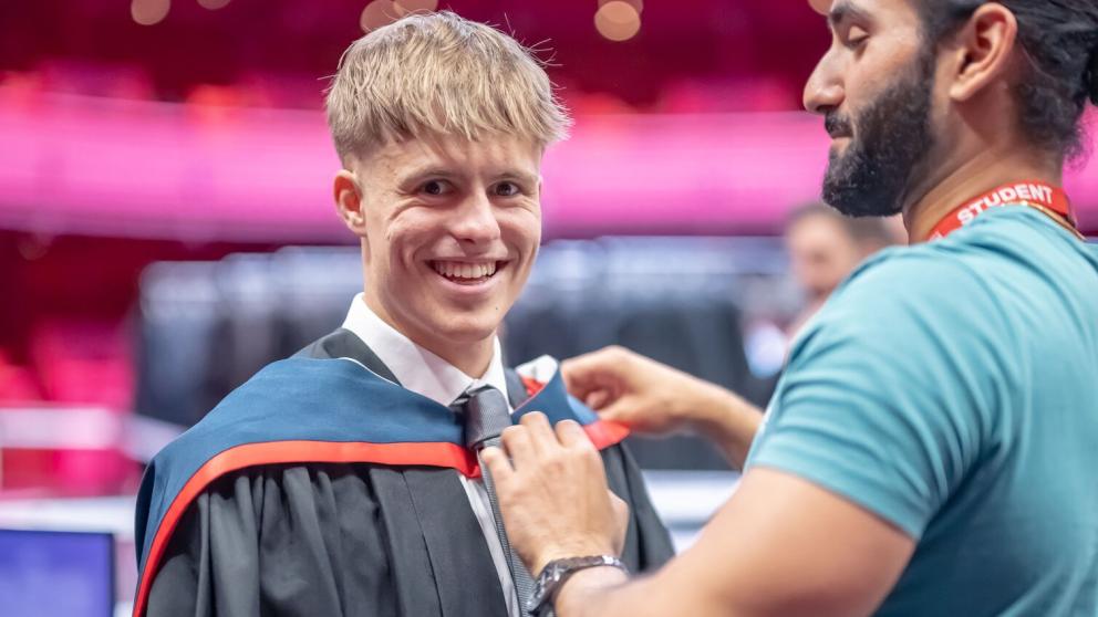 Graduate smiles at the camera as a student ambassador helps them put their graduation gown over their suit. The gown is black and the hood is red and dark blue.