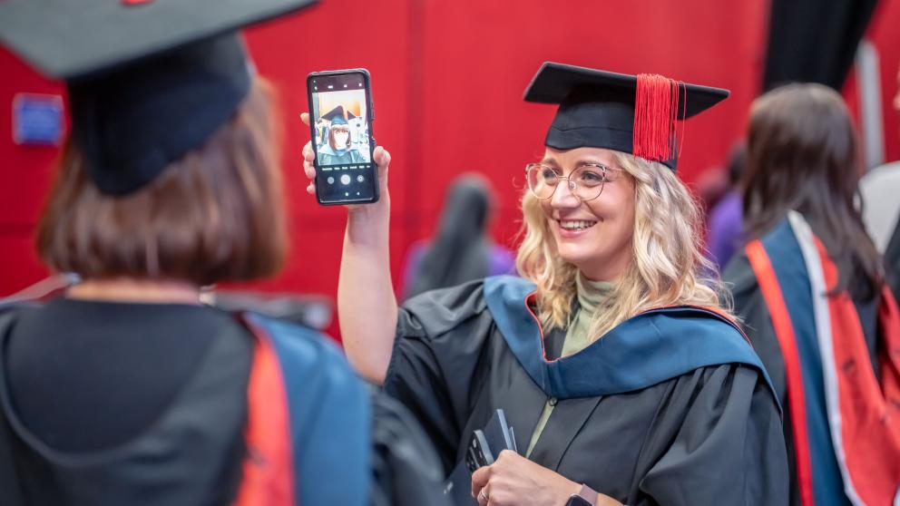 A graduate in a black graduation gown and blue and red hood, holds up their phone for another graduate stood across from them in the same graduation attire. Both wear black mortar board hats with red tassels
