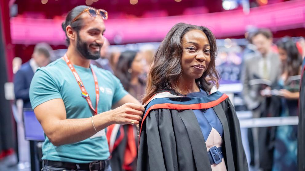 A graduate smiles at the camera while wearing a black gown over their blue and white dress. A student ambassador adjusts their graduation hood which is blue and red from behind them.