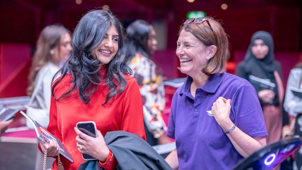 A graduate in a red dress holds a Graduation brochure, graduation ticket and their black robe as they laugh with a staff member in a purple polo shirt.
