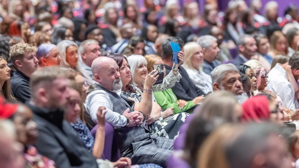 Guests in the audience of Graduation sit in purple seats and look at the stage. A couple in the middle look at the stage with emotional gazes.