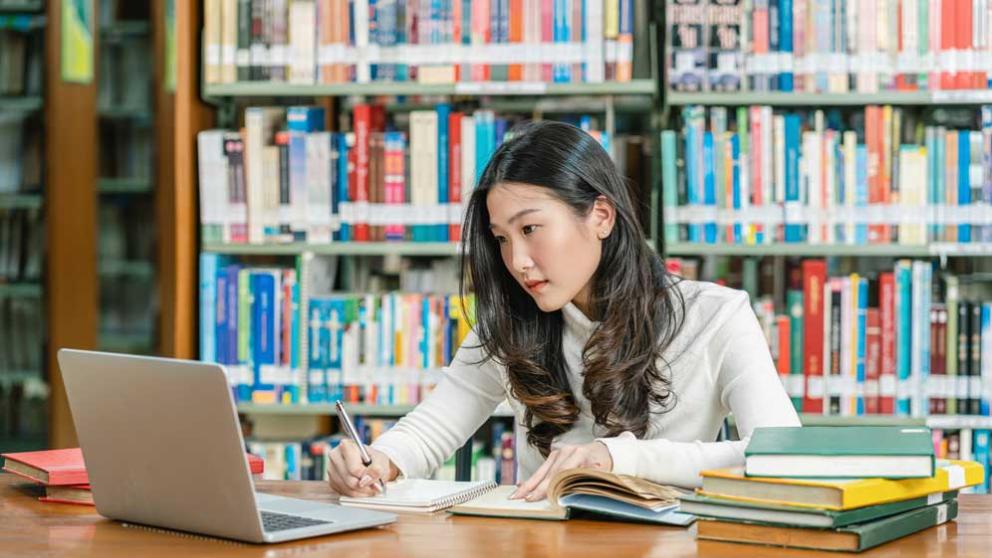 Student in a library studying with books and a laptop