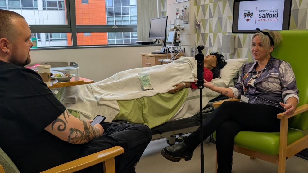 A man and a woman sat in a hospital simulation suite, filming a podcast with a simulation dummy in the background