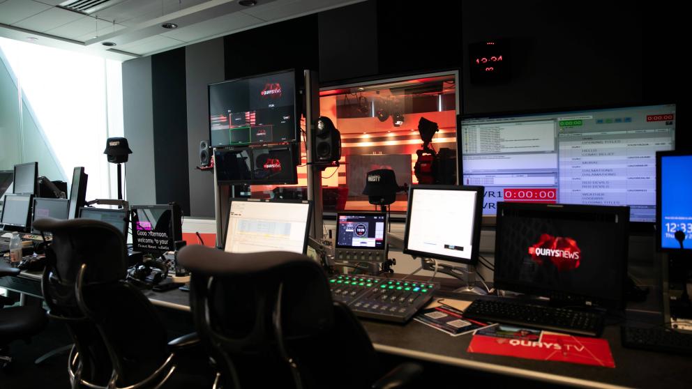 News studio gallery with multiple computer monitors and a large screen showing a presenter