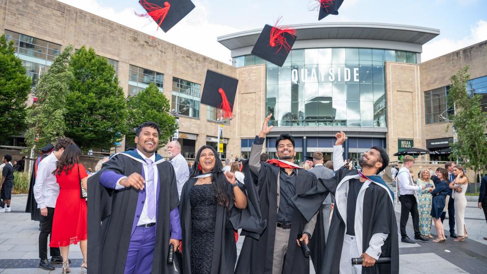 Graduates wearing caps and gowns celebrate outdoors, they smile at the camera and throw their caps into the air above a crowd of people. Trees surround the area and the sky is partly cloudy.