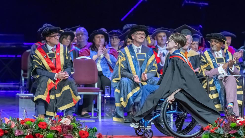 A graduate in a wheelchair wearing academic robes crosses the front of the stage academics behind them applaud. Red and green floral arrangements line the stage.