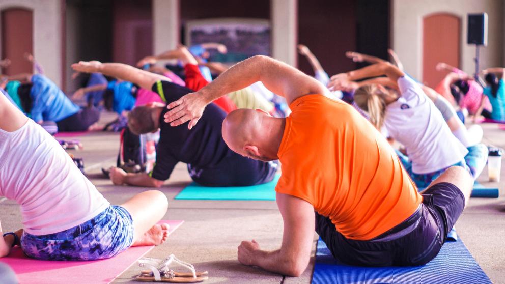 A diverse group of people sit on yoga mats facing away from the camera. They are all leaning to one side, with one arm raised over their heads as they stretch and reach sideways.