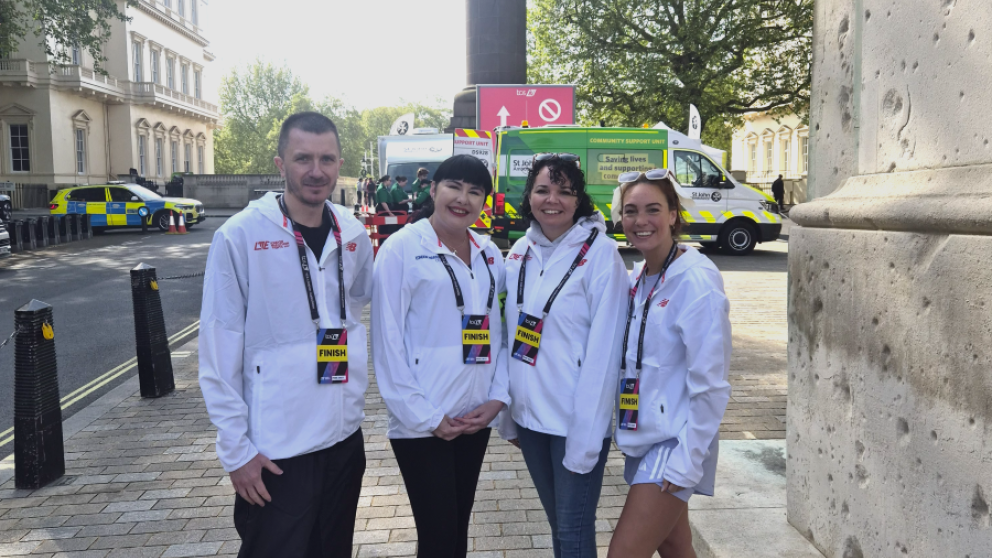 A group of four podiatry students and staff smile at the camera. They are stood on the street in front of a number of support vehicles and wear official London Marathon branded jackets and lanyards.