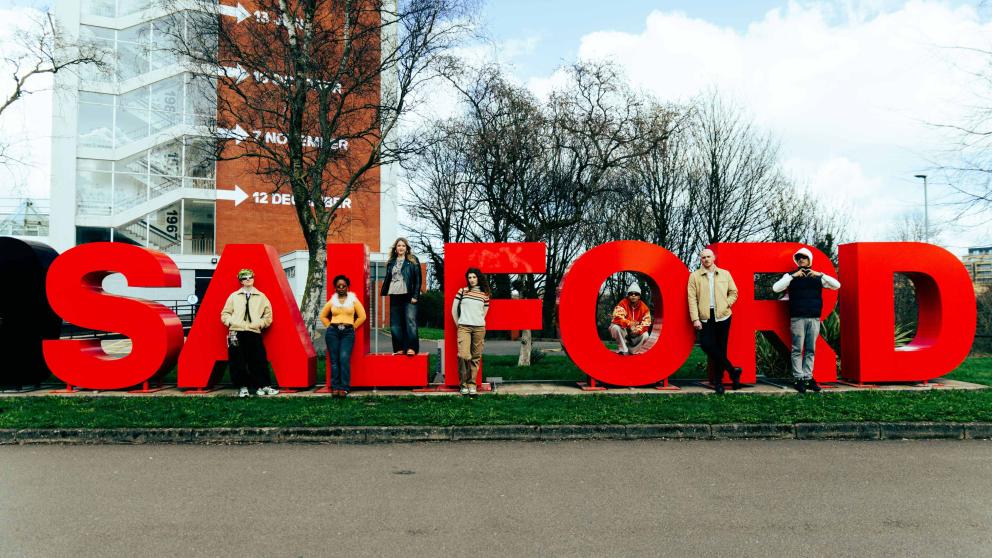 A group of six people pose in front of the Salford sign