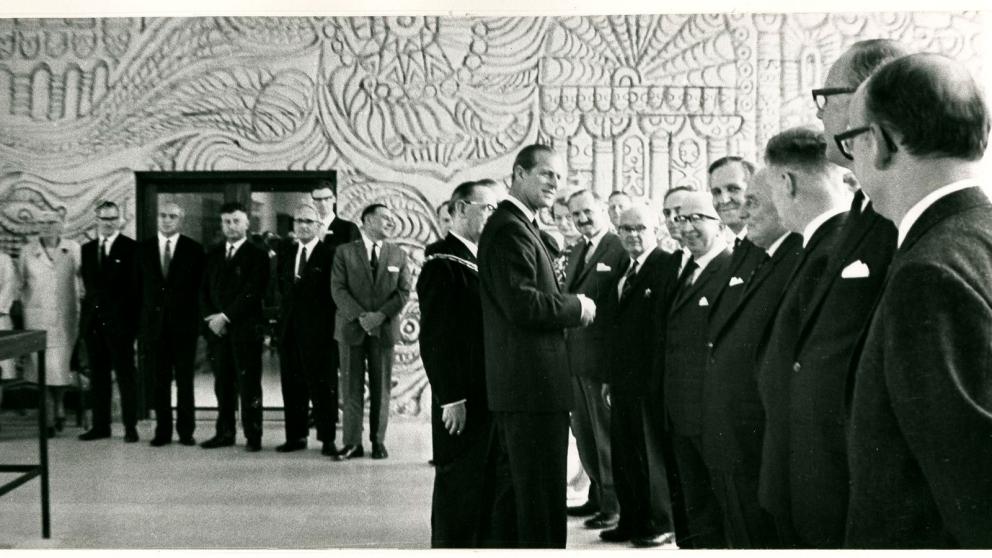 A formal reception in a large decorative hall. Prince Philip is among a line of men in dark suits greet and converse with a tall man at the center, while additional attendees stand in the background near a doorway. Intricate mural artwork covers the wall behind them.