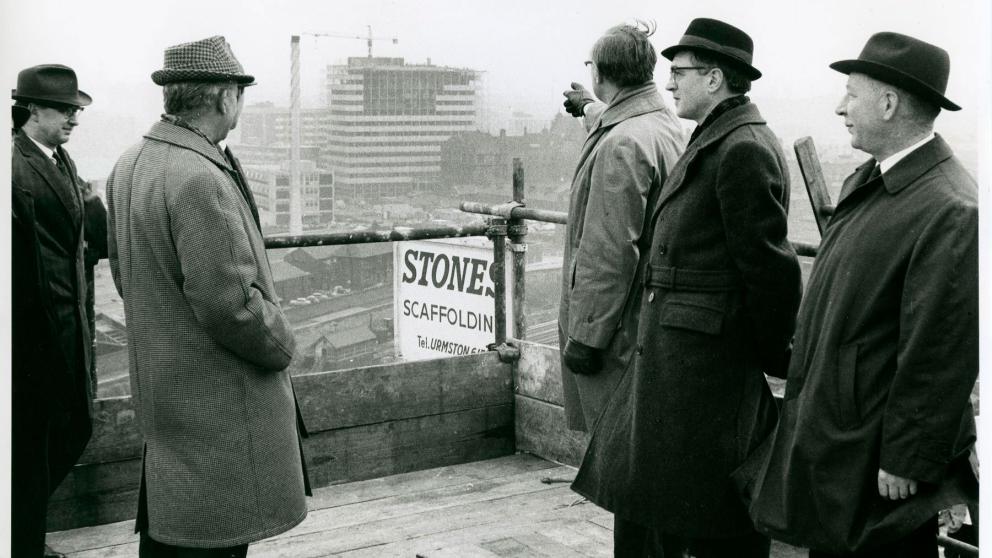 Five men wearing overcoats and hats standing on a wooden scaffolding platform overlooking a city construction site. One man points toward a multi-story building under construction in the distance, while the others look on.