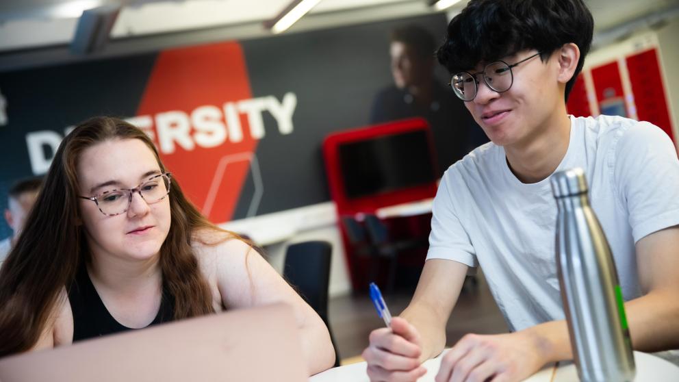 two sbs students sat smiling, one with a laptop and one with a pen and notebook and waterbottle