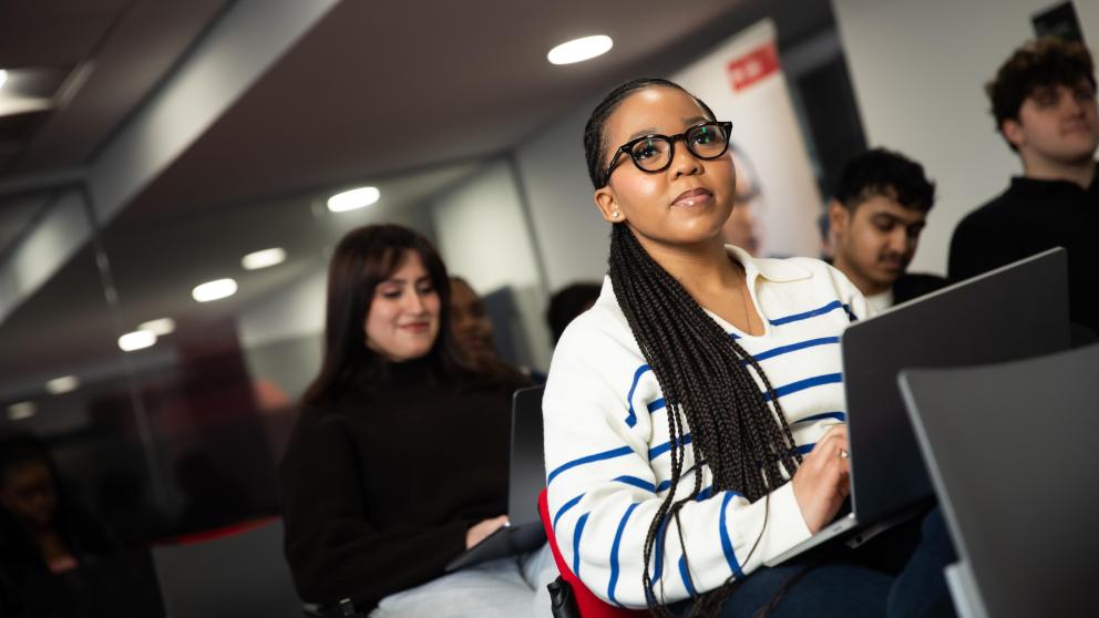 sbs student in focus sat looking ahead in business lecture, other students behind out of focus