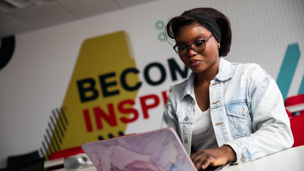 sbs student looking down at laptop in front of wall that reads 'become inspired'