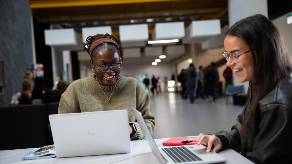 two students sat at a table smiling on their laptops