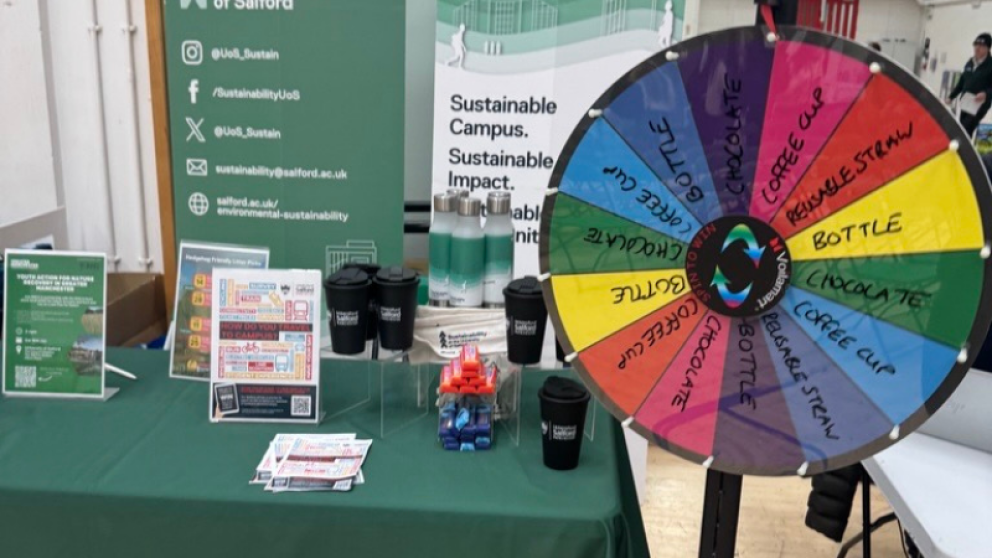 A photograph of a table with green tablecloth and various promotional items such as a bottle, coffee cups and posters with a rainbow coloured spinning wheel with prizes in the foreground