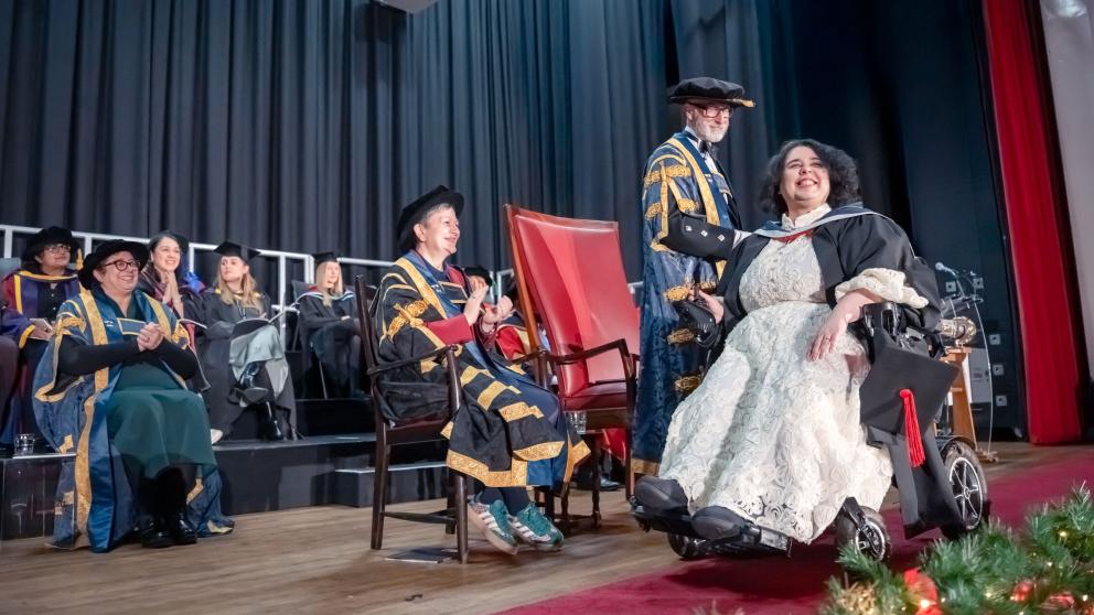 A graduate in academic dress sits in a powered wheelchair on a stage during a graduation ceremony, holding a mortarboard with a red tassel. University officials in blue and gold ceremonial robes are seated and standing nearby, while other graduates and staff are visible in the background. The stage is framed by dark curtains and red carpeting, with festive greenery along the front edge.