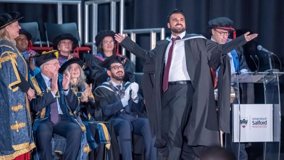Graduate in academic dress celebrating on stage with faculty members, University of Salford