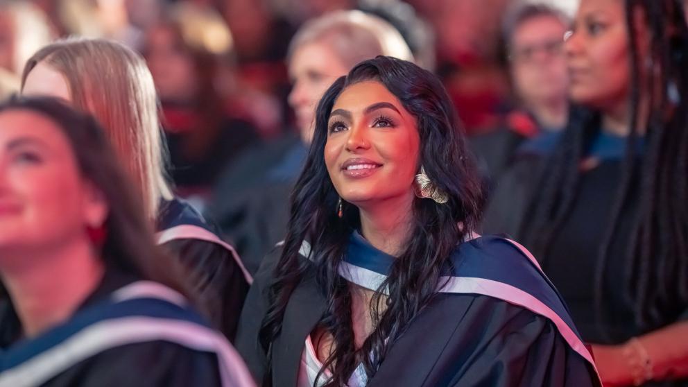A graduate wearing a black academic gown with a blue-and-white hood is seated among other graduates at a graduation ceremony. The graduate has long dark hair and large metallic earrings and is looking upward. The background shows rows of attendees and graduates in soft focus, illuminated by warm red and pink stage lighting, creating a celebratory atmosphere.
