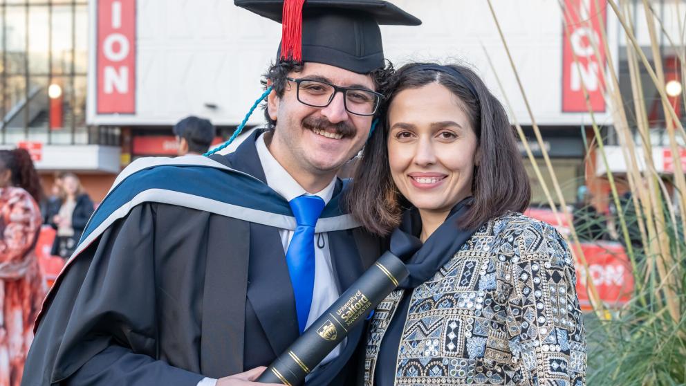 Graduate in academic dress holding a degree certificate alongside another person in front of Maxwell Building, University of Salford