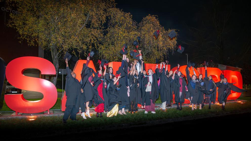 A large group of graduates in black academic gowns gather outdoors at night in front of illuminated red letters spelling “SALFORD.” Many are throwing their mortarboards into the air while standing on a grassy area beside a path. Trees with autumn foliage form the backdrop, and the scene is brightly lit, highlighting the graduates and the University of Salford landmark sign during a graduation celebration.
