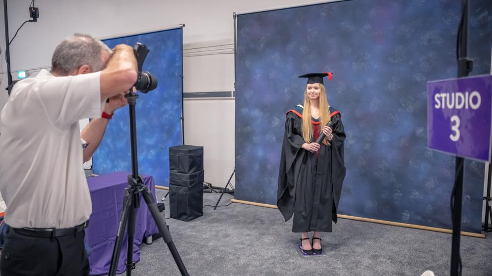 A graduation photography studio setup with a photographer taking a portrait of a graduate standing in front of a blue mottled backdrop. The graduate wears a black academic gown, mortarboard with a red tassel, and red-trimmed hood, and holds a diploma tube. A purple sign reading “STUDIO 3” is visible on the right, with photography equipment and backdrop stands positioned around the studio space.