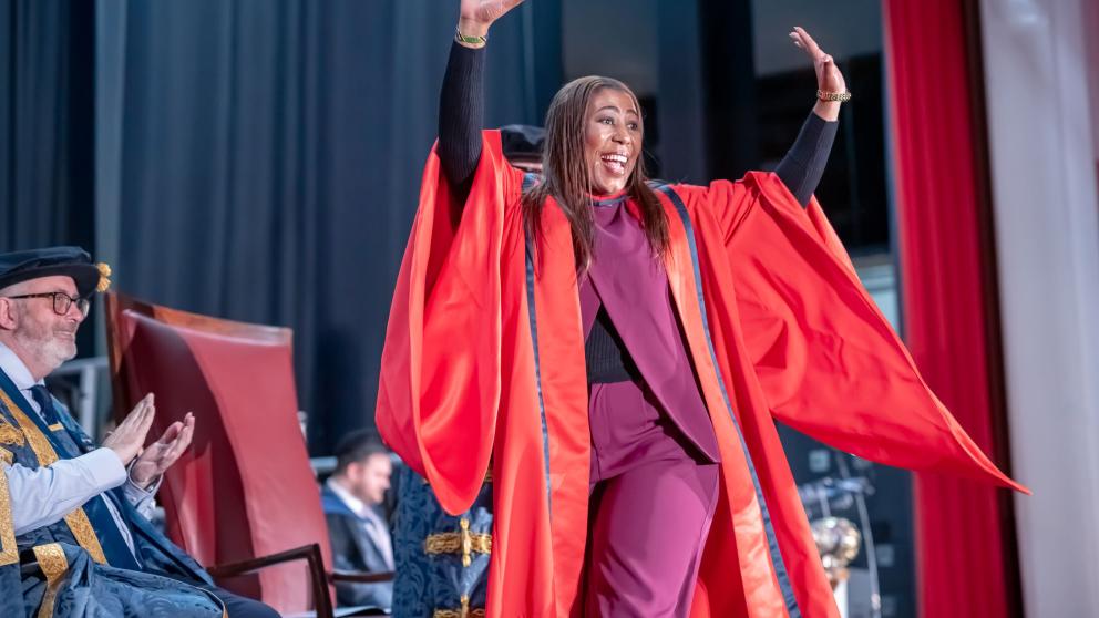 A person wearing a bright red academic robe raises both arms while crossing a graduation stage. Seated academic officials in ceremonial robes applaud from the side of the stage, while dark curtains and red drapes form the backdrop. The image captures a celebratory moment during a graduation ceremony.