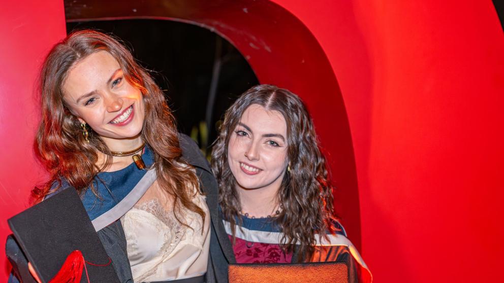 Two graduates in academic gowns pose together in front of a large red sculptural backdrop, each holding a mortarboard with a red tassel. One graduate wears a light-coloured outfit beneath a black gown, while the other wears a patterned outfit with academic hood colours visible. The photograph is brightly lit, with the vivid red background creating a celebratory graduation setting.