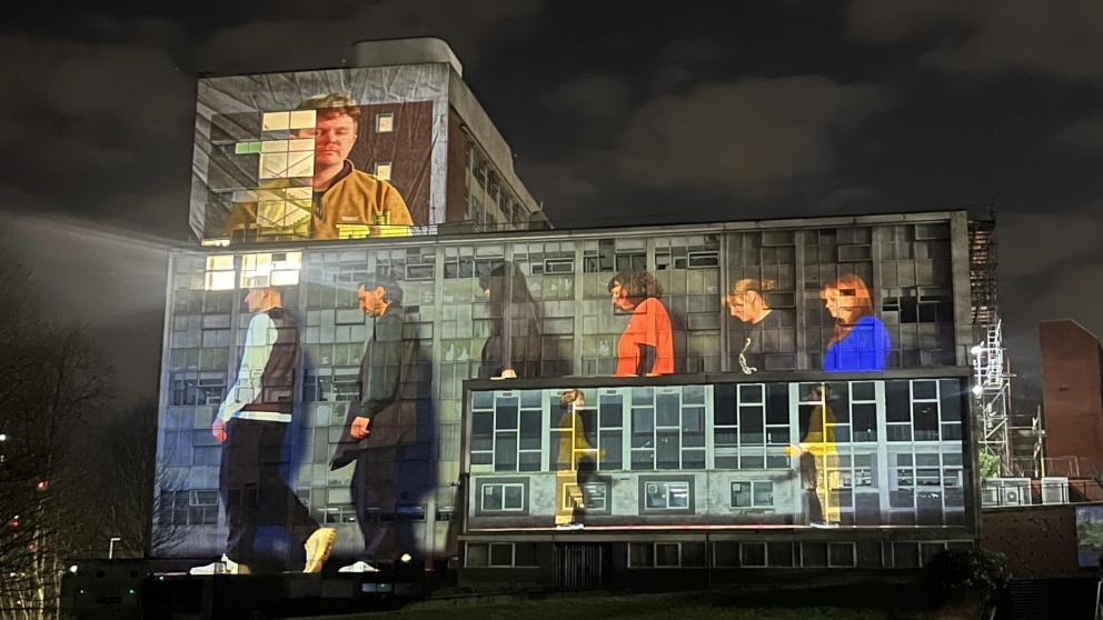 Images of six people walking with their heads down are projected onto the side of Maxwell building against a night sky
