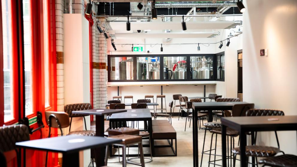 The interior of The Old Fire Station. Industrial looking decor with chairs grouped around tables. You can see the brewing equipment in a back room through large glass windows.
