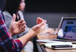 Two people in a meeting, with one person's hands gesturing near a laptop displaying graphs or data on the screen