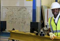 A student stood in our Heavy Structures lab with his hands on a metal beam and smiling while wearing a yellow hi-vis vest and a white hard hat
