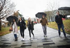 Group of 5 students walking along the University broadwalk in the rain, holding umbrellas.
