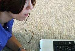 A student works on a laptop while holding their reading glasses in their mouth