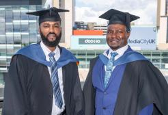 Two men in suits and graduation attire posing for photograph. Big billboards behind them read 'Dock 10' and 'Media City UK' 