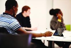Three students sit around a table in a classroom looking at something out of frame. One has an iPad.