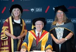 Two women stand next to a man, all in graduation attire in front of wall with University of Salford logo 