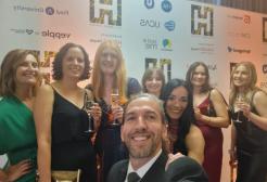 Six women in dresses and a man in a suit pose for a photograph in front of a wall with different logos on it. 