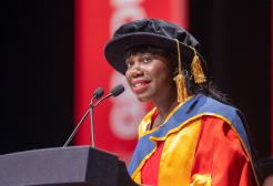 Dr Sally Penni giving a speech in red, yellow and blue graduation attire on stage in front of a large red banner